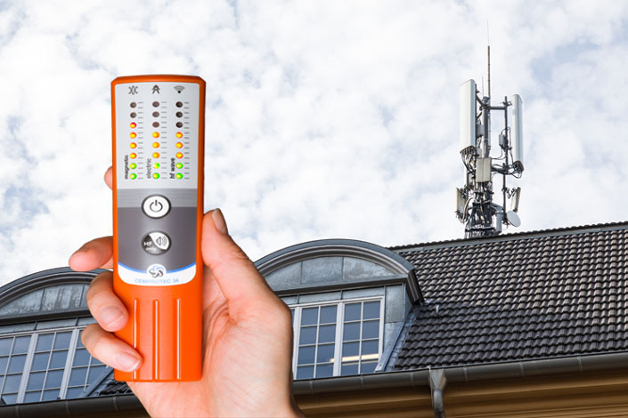 Un appareil de mesure de l'électrosmog Cemprotec 34 devant une maison avec une antenne de téléphone portable.