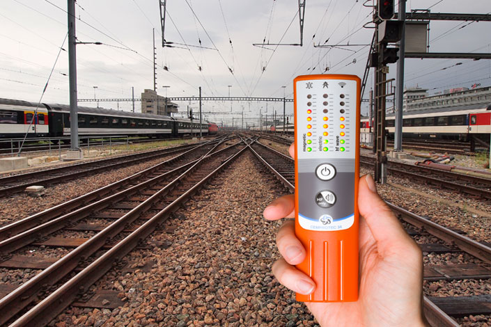 Eine Person hält ein Cemprotec 34 Hochleistungs-Elektrosmog-Messgerät vor einem Bahngleis.