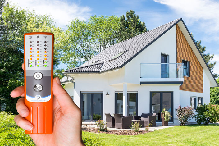 Une personne tient un appareil de mesure de l'électrosmog Cemprotec 34 haute performance devant une maison.