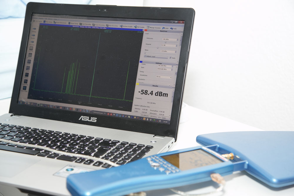 Un ordinateur portable sur lequel s'affiche un programme d'analyse de signal avec une valeur de -58,4 dBm, à côté d'un appareil de mesure manuel bleu avec une antenne connectée.